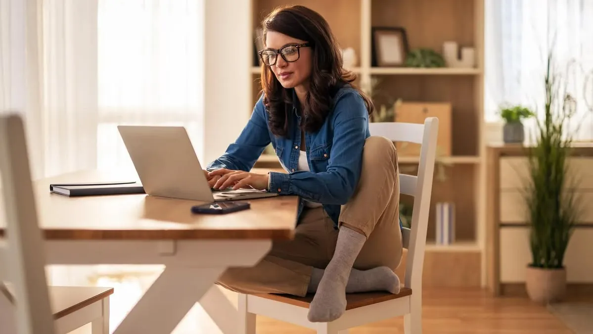 A woman in a denim shirt working on a laptop at a wooden dining table in a bright, calm room